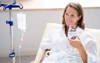 lady in medical clinic with IV drip drinking a glass of water, smiling